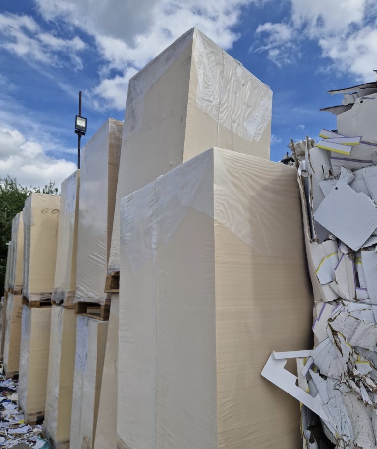 Tall plastic-wrapped stacks of paper sheets on pallets beside a bale of printed card